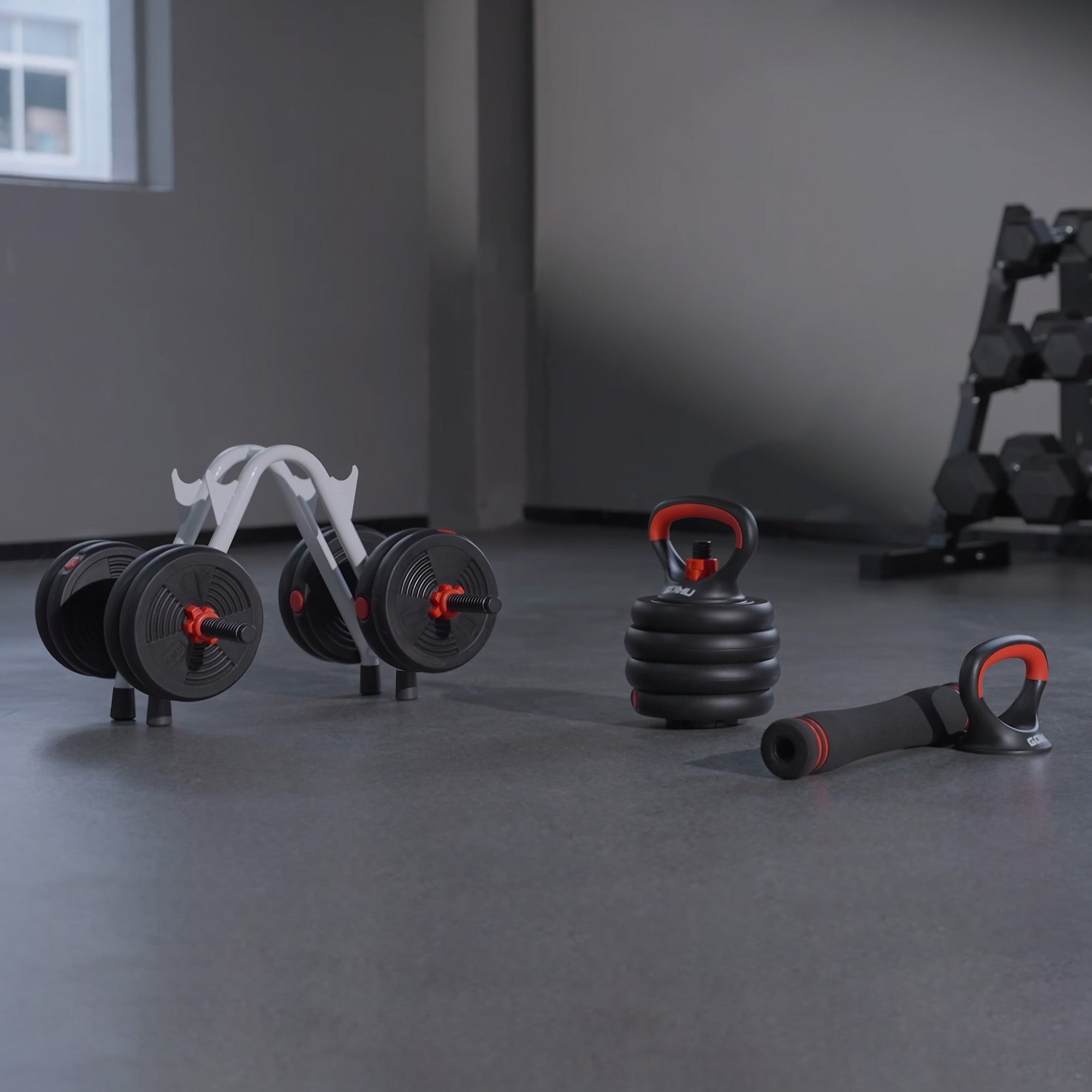 Set of adjustable dumbbells and kettlebells on a gray floor with a rack in the background.