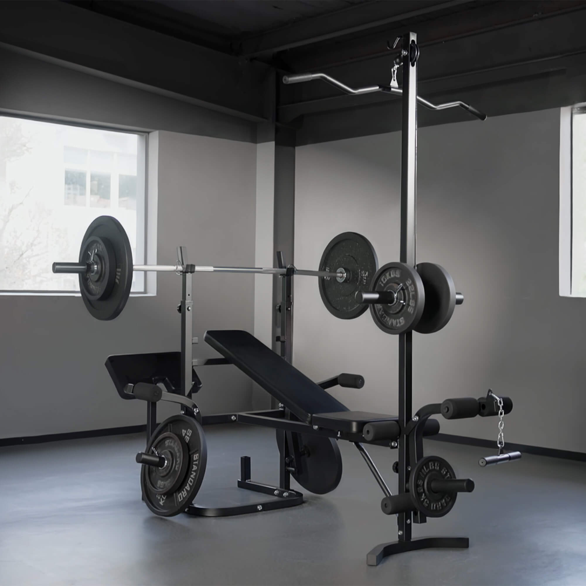 Gym equipment including a barbell, weight plates, and a bench in a room with gray walls and a window.
