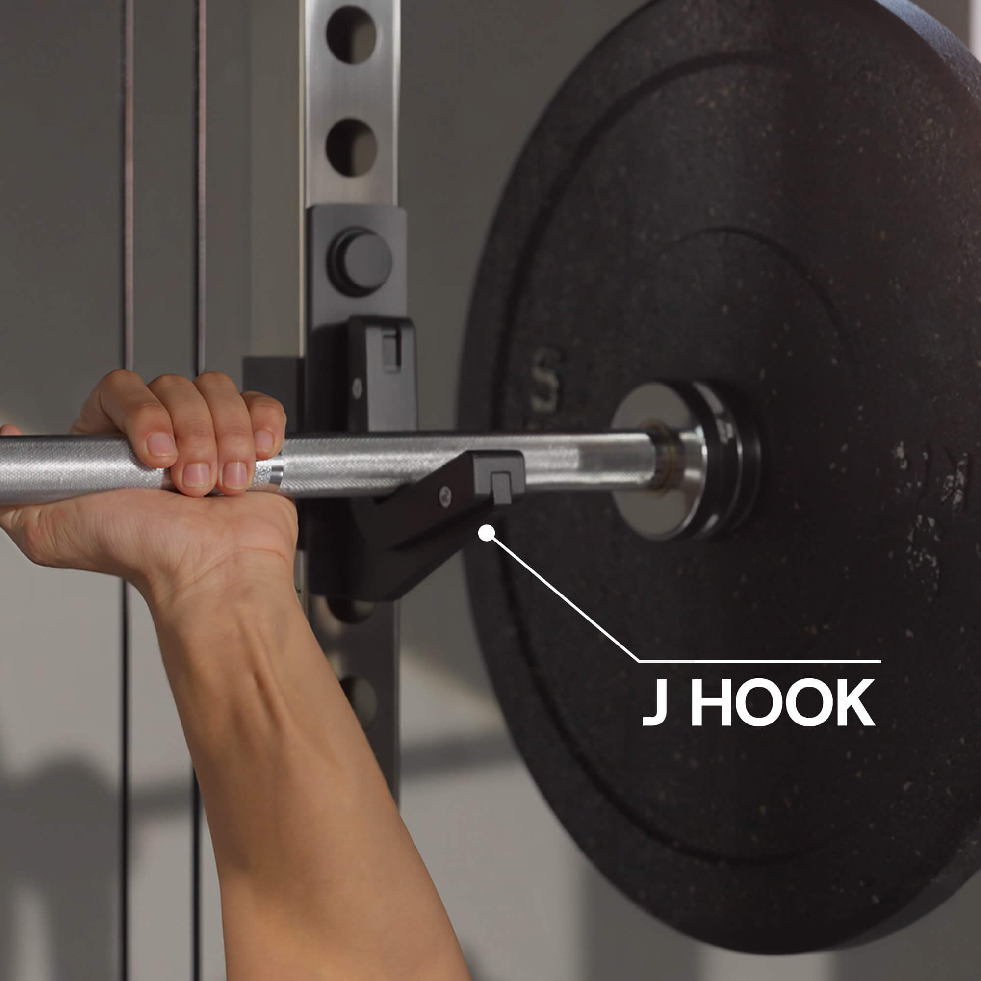 Hand holding a barbell with a J hook labeled on a gym background.#color_black